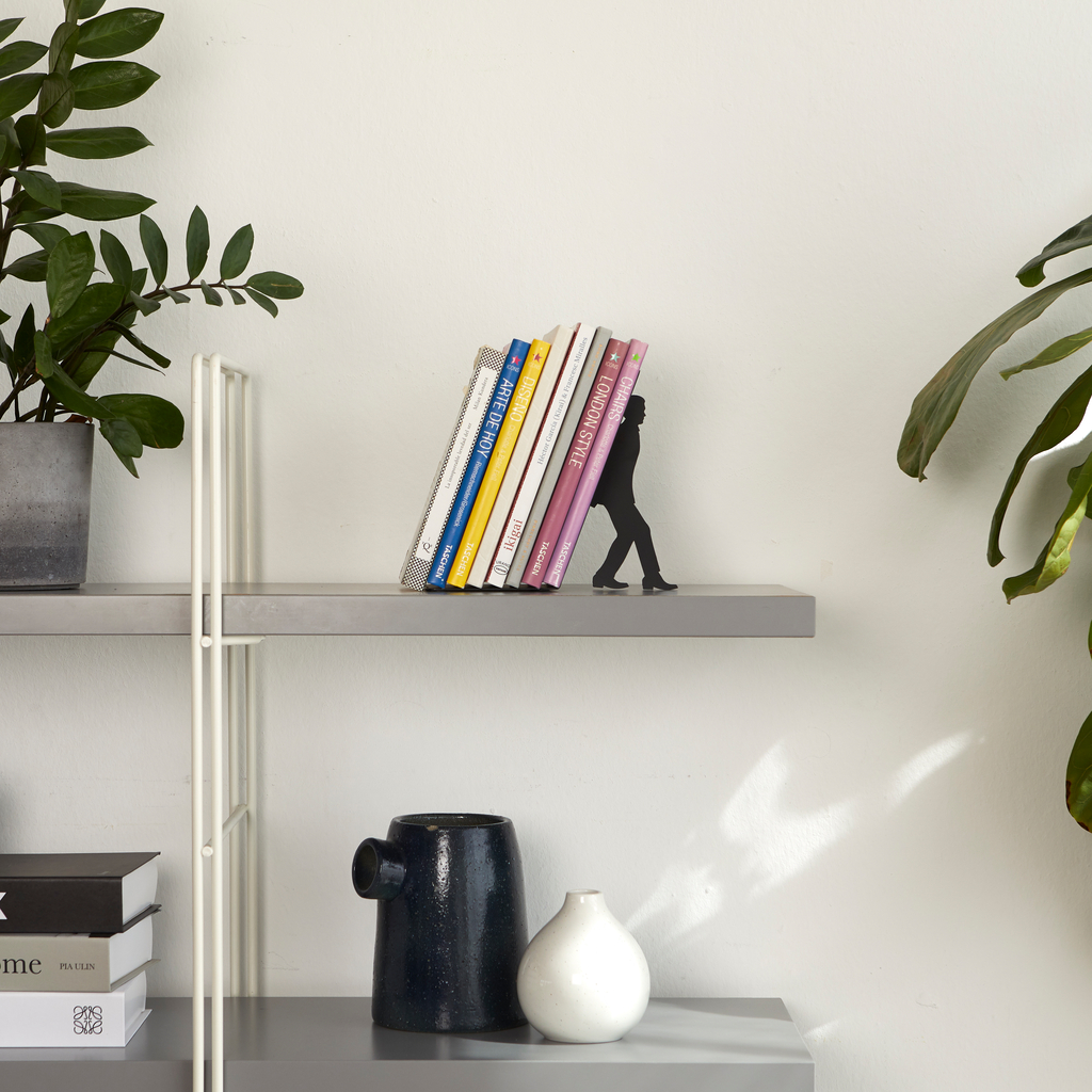 Zwarte Balvi boekensteun met een rij boeken op een moderne wandplank tussen groene planten
